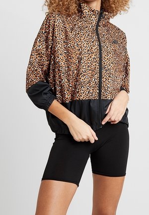 Vrouw draagt een jas met luipaardprint en zwarte ritssluiting met elastische manchetten en zwarte strakke fietsshorts.