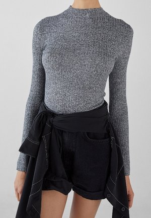 Mujer con un suéter gris acanalado de manga larga y pantalones cortos vaqueros negros con una chaqueta negra atada alrededor de la cintura.