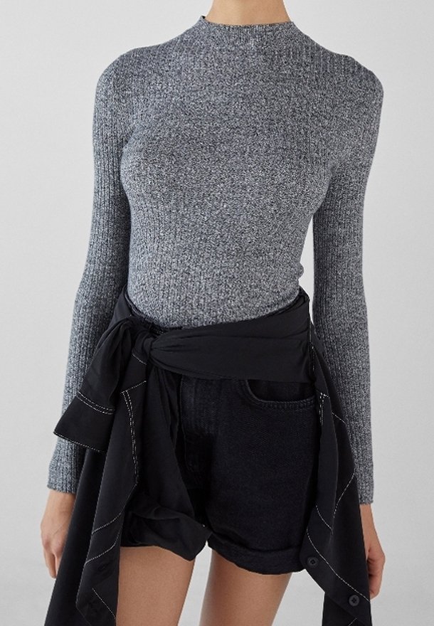 Femme portant un pull gris côtelé à manches longues et un short en jean noir avec une veste noire nouée autour de la taille.
