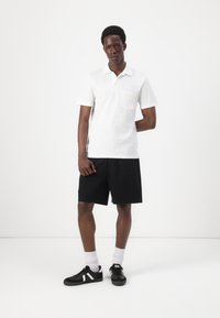 Hvit kortermet polo skjorte med brystlomme, parret med svarte shorts. Modellen har på seg svarte sko med hvite detaljer og hvite sokker.