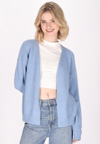 Cardigan lavorato a maglia di un azzurro chiaro, design aperto nella parte anteriore, texture morbida, polsini a costine, indossato sopra una maglietta bianca, abbinato a jeans a vita alta.