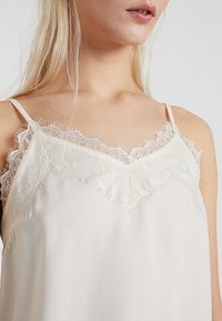 Krémová tílko s jemným krajkovým lemem kolem výstřihu a nastavitelnými tenkými ramínky. Měkká, lehká tkanina s hladkým povrchem.