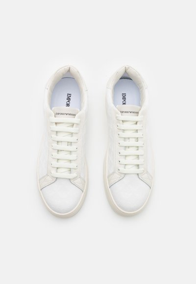 White sneakers with a smooth synthetic upper, flat laces, and a textured design. Features a cushioned insole and round rubber outsole.
