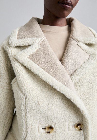 Crème-kleurige dubbelbreasted jas met een fuzzy textuur, voorzien van een contrasterende beige kraag en twee grote knopen voor sluiting.