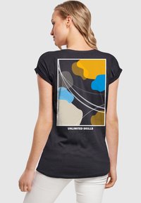 Zwarte T-shirt met korte mouwen, voorzien van een abstract ontwerp in oranje, blauw, geel en beige met de tekst "ONBEPERKTE VAARDIGHEDEN" onderaan.