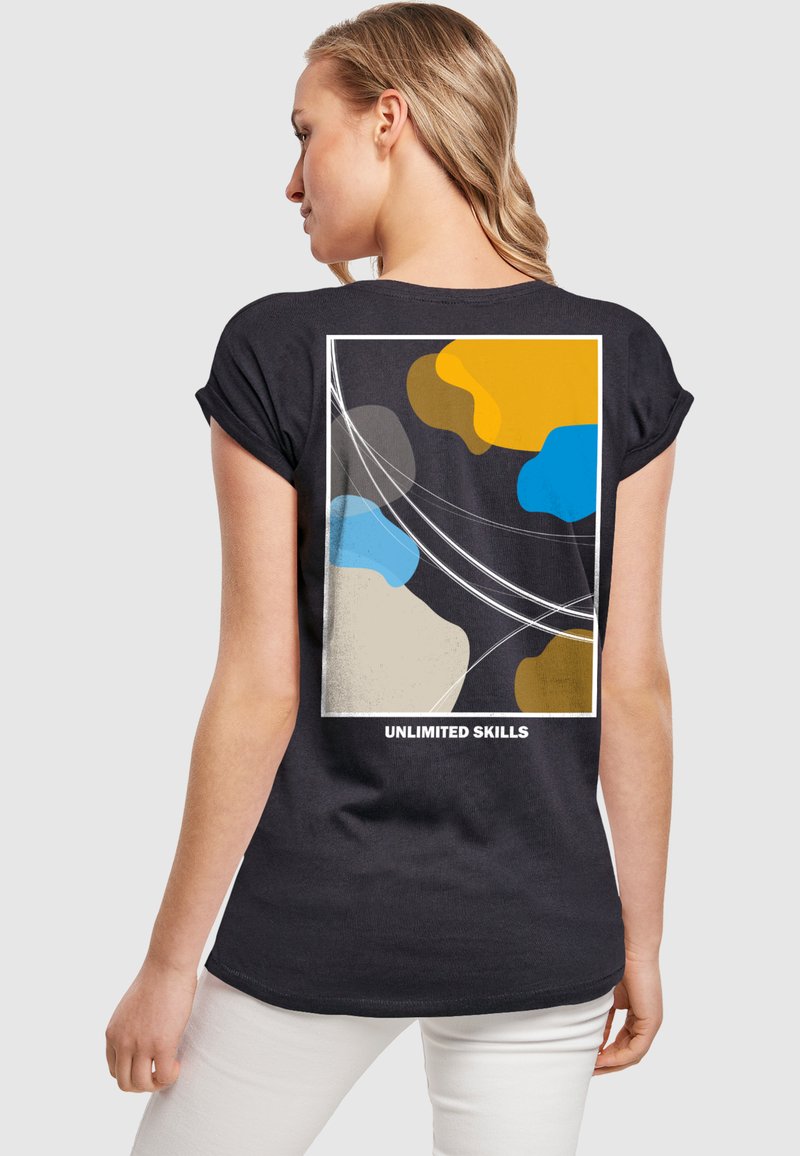 Zwarte T-shirt met korte mouwen, voorzien van een abstract ontwerp in oranje, blauw, geel en beige met de tekst "ONBEPERKTE VAARDIGHEDEN" onderaan.