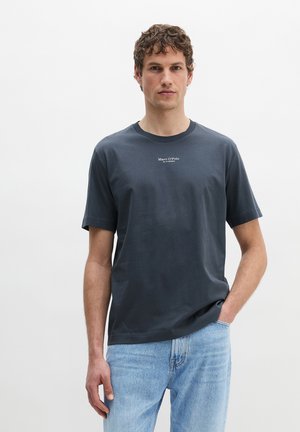 Mężczyzna w ciemnoszarej koszulce Marc O’Polo i jasnoniebieskich dżinsach, z jedną ręką w kieszeni, stoi na jednolitym białym tle.