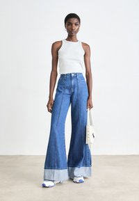 Rozšířené džíny z denimu s barevným blokem v světle modré a tmavě modré barvě, vysoký pas, kontrastní švy, spárované se šedým topem na ramínka a bílou kabelkou.