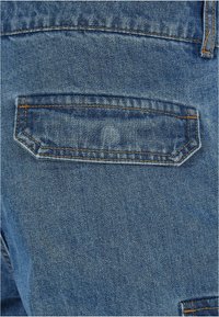 Vaqueros de mezclilla con un bolsillo trasero con un diseño de parche, en un lavado azul medio con costuras naranjas en un tejido texturizado.