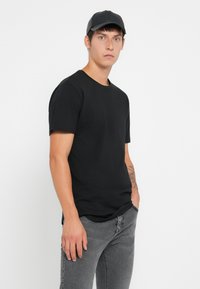Hombre joven con un tatuaje en el antebrazo izquierdo vestido con gorra negra, camiseta negra y jeans grises, frente a un fondo blanco liso.