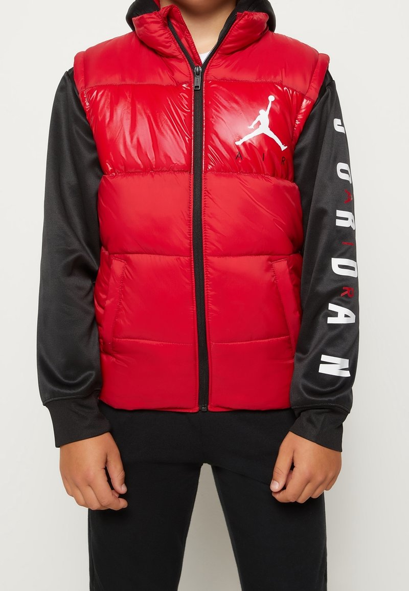 Czerwona kamizelka puchowa z błyszczącym wykończeniem, z zamkiem z przodu i kapturem. Na piersi białe logo Jordan, czarne rękawy z białym napisem „JORDAN”.