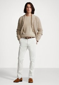 Beżowy dzianinowy kardigan na jasnym swetrze, zestawiony z białymi spodniami i brązowymi mokasynami. Model stoi z rękami w kieszeniach na jednolitym tle.