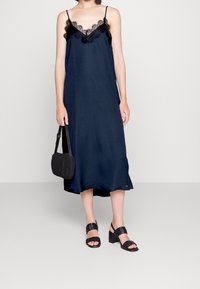 Mörkblå, texturerad slipklänning med svarta spetsdetaljer, kombinerad med en svart crossbody-väska och klumpiga svarta sandaler.