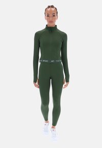 Olívazöld sportos szett cipzáros dzsekivel és hozzáillő leggingsszel, márkás derékpánttal és hosszú ujjakkal. Fehér sportcipő.