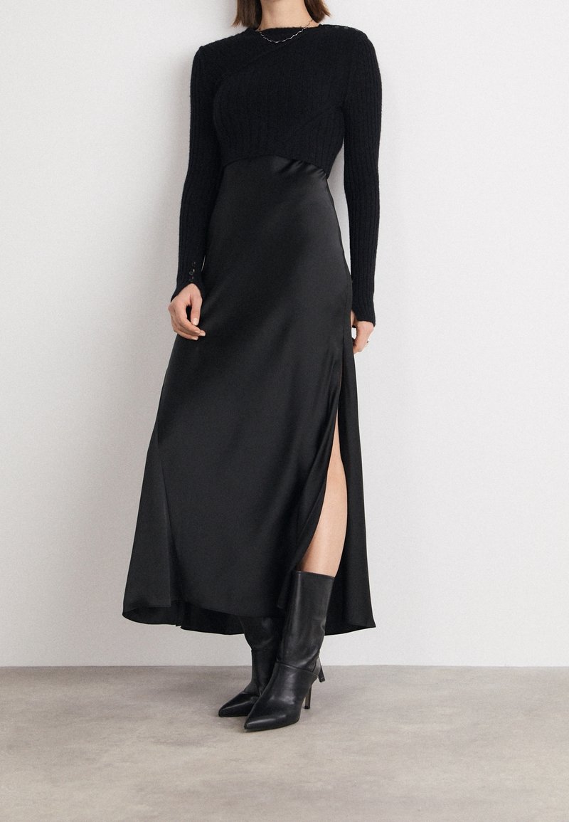 Un haut noir en maille à manches longues associé à une robe maxi en satin noir, avec une fente sur le côté et des bottes à talons noires, créant un look superposé.