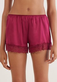 Bordeaux satijnen shorts met een elastische tailleband. Afgewerkt met een golvende kantafwerking aan de zoom, waardoor textuur en ontwerpdetails worden benadrukt.