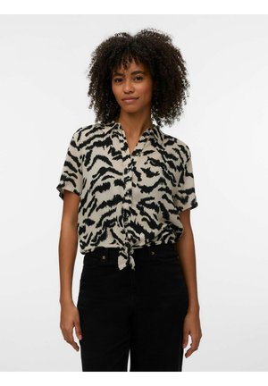 Mujer con cabello rizado que lleva una camisa de manga corta abotonada con rayas de tigre en beige y negro, atada a la cintura, y pantalones negros.