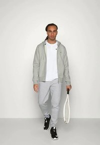 Egy férfi szürke kapucnis pulóverben, fehér pólóban, szürke melegítőnadrágban és fekete sportcipőben sétál, miközben egy fehér teniszütőt tart egy egyszínű háttér előtt.