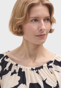 Lehká blůza bez ramínek se smetanovým pozadím a tmavomodrými abstraktními květinovými vzory. Měkká látka s nařaseným výstřihem.