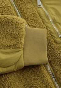 Povečava rokava olive zelene jakne z pletenim manšetom, teksturirano flis tkanino in srebrnimi zadrgami na tkaninskih panelih.