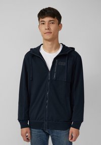 Marineblauwe ritskap met een zachte stof, voorzien van een voorzak, een trekkoordcapuchon en geribde manchetten. Draag het over een wit T-shirt.
