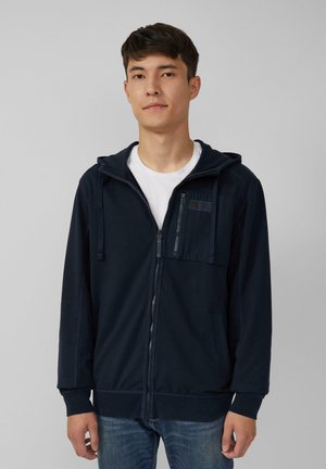 Navy zip-up hoodie made of soft fabric, featuring a front pocket, drawstring hood, and ribbed cuffs. Worn over a white t-shirt.