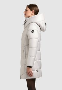 Lys grå pufferjakke med hætte, der har en quiltet design, lynlåse og logo detaljer. Lange ærmer med sorte manchetter og sidelommer.