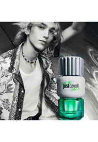 Jeune personne portant une chemise à motifs et un collier de coquillages, posant à côté d'un flacon de parfum vert et argenté « Just Cavalli Magic ».