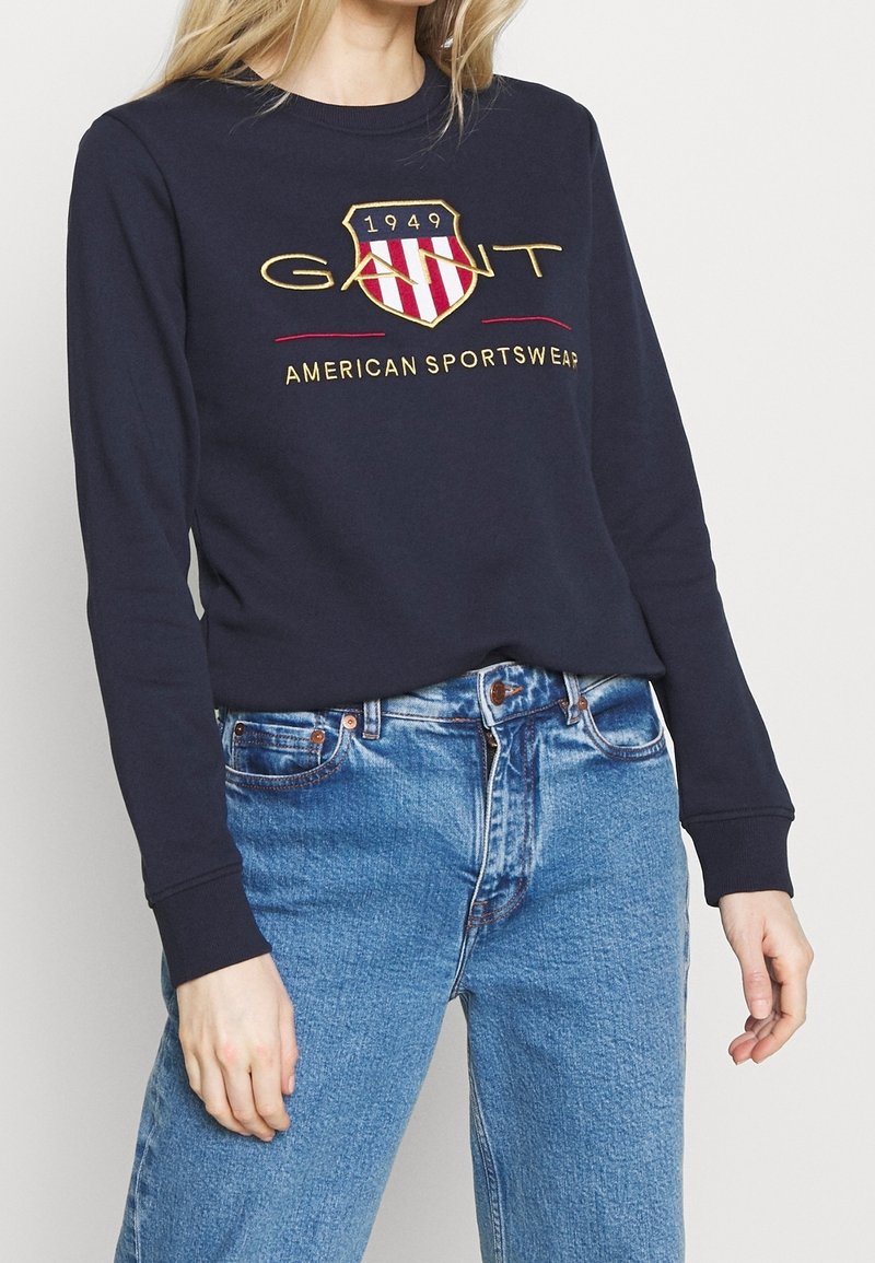 Femme portant un sweat-shirt bleu marine avec un logo écusson GANT rouge et or et le texte « American Sportswear », associé à un jean bleu clair.