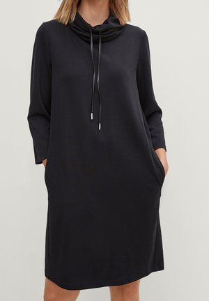 Femme portant une robe noire à manches longues, longueur genou, avec un col bénitier ample et des cordons, mains dans les poches latérales.