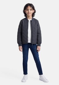 Bērns ar plecu garuma tumšiem matiem, valkājot melnu dūnu jaku, baltu kreklu, tumšas džinsas, baltas zeķes un baltas sporta kurpes, stāvot pret baltu fonu.