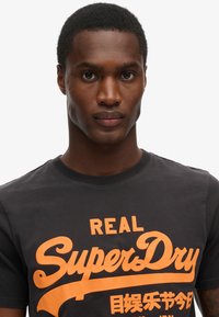 Camiseta de algodón negra con un audaz logotipo y texto naranja de "SuperDry". Cuello redondo, mangas cortas, con una textura suave y un ajuste casual.