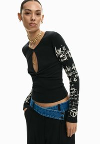 Zwart top met lange mouwen, een sleutelgat aan de halslijn, geribbelde textuur en witte schreefdetails op de mouwen. Gecombineerd met een tailleband met blauwe denimaccenten.