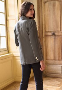 Woman in gray blazer and dark pants looking back standing near window and wooden door in a bright room with parquet floor.