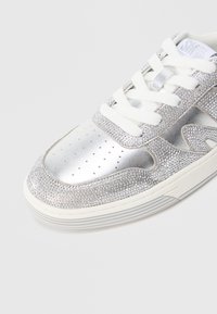 Ασημί αθλητικό παπούτσι με υφή στο πάνω μέρος και μικρές στρας, διάτρητο στα δάχτυλα και λευκά κορδόνια. Λαστιχένια σόλα με ασημί λεπτομέρειες.