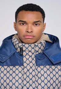 Giacca imbottita con una parte superiore blu navy e una parte inferiore beige caratterizzata da un motivo a rombi. Chiusura con zip e design del colletto alto.