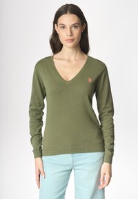 Donna con lunghi capelli castano scuro, che indossa un maglione verde oliva a V e jeans azzurri, in piedi contro uno sfondo semplice.