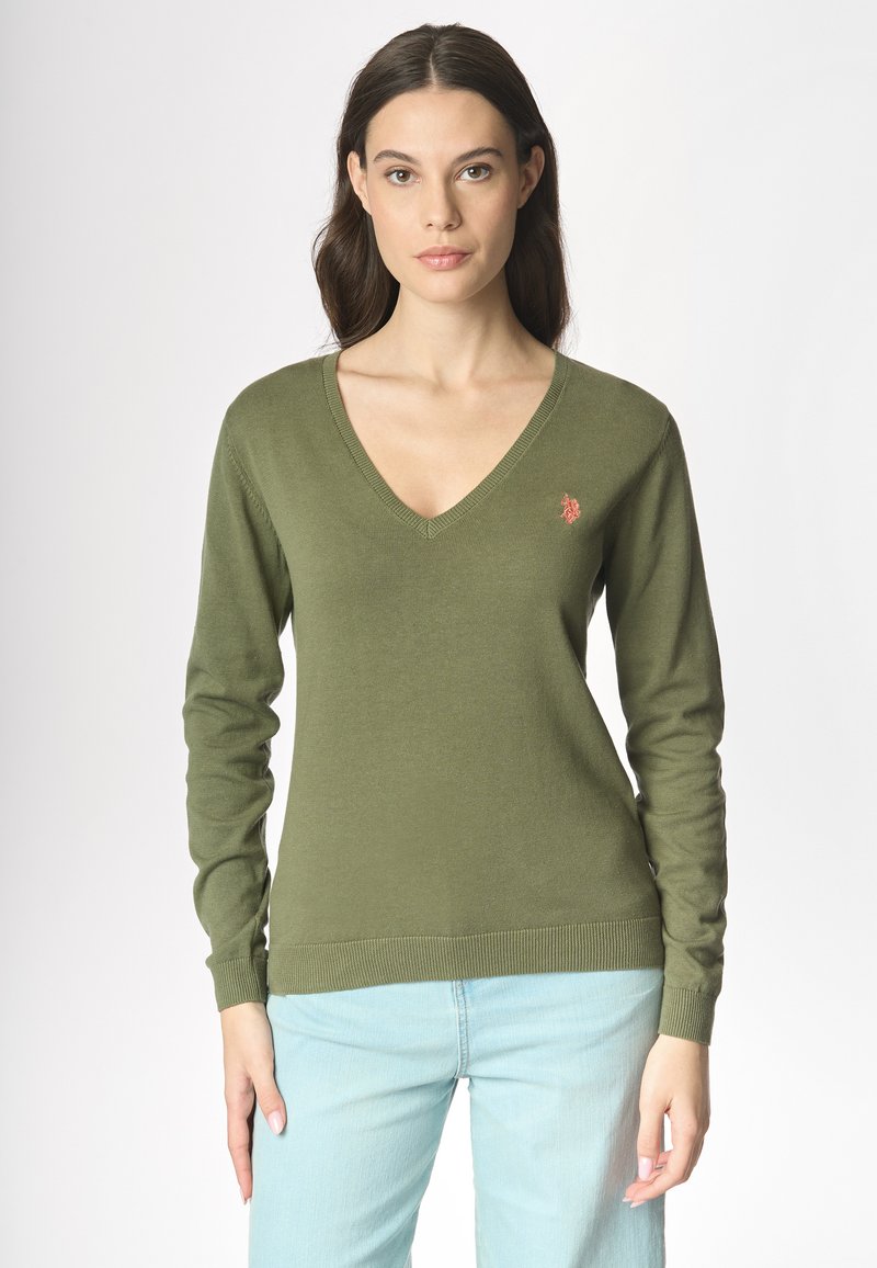 Donna con lunghi capelli castano scuro, che indossa un maglione verde oliva a V e jeans azzurri, in piedi contro uno sfondo semplice.