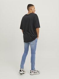 Muškarac stoji s rukama u džepovima, odjeven u crnu oversized majicu, svijetloplave uske traperice i bijele tenisice, okrenut leđima prema jednostavnoj pozadini.