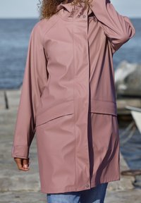 Personne portant une veste imperméable rose poudré à boutons-pression au bord de la mer, debout sur une jetée en béton près de l'eau et des rochers.