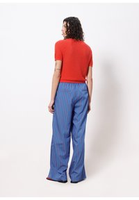 Rotes gestricktes Kurzarmoberteil und blaue weit geschnittene Hose mit vertikalen roten Streifen. Die Hose hat eine Gesäßtasche und ist leicht ausgestellt.