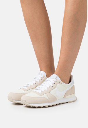 Hvide og beige Nike løbesko med snørebånd, båret på bare ben mod en hvid baggrund.