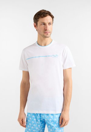 Biała koszulka z krótkim rękawem, z jasnoniebieskim obramowaniem kołnierzyka i poziomą niebieską linią na wysokości klatki piersiowej; po lewej stronie znajduje się logo w postaci palmy.