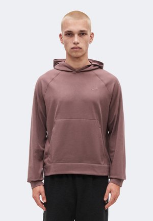 Jeune personne à la tête rasée portant un sweat à capuche mauve et un pantalon noir, debout face à l’avant sur un fond uni.