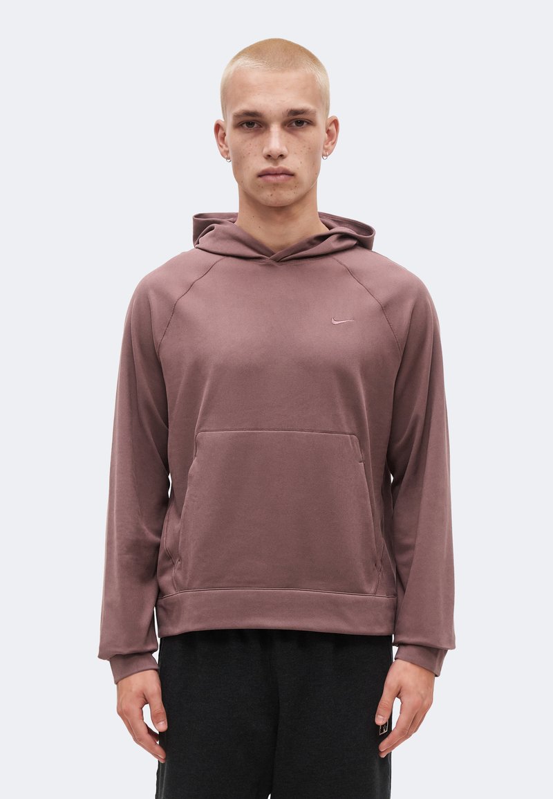 Νεαρό άτομο με ξυρισμένο κεφάλι, που φοράει μπουφάν χρώματος μωβ και μαύρο παντελόνι, στέκεται με το πρόσωπο προς τα εμπρός σε απλό φόντο.