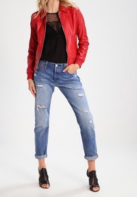Rote Leder-Bomberjacke mit gerippten Ärmelbündchen, darunter ein schwarzes, durchsichtiges Oberteil, ausgewaschene blaue Jeans und offene schwarze High Heels.