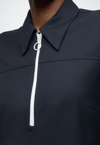 Μπλε ναυτικό τοπ με φερμουάρ μέχρι τη μέση, με δομημένο γιακά, αντίθετο λευκό φερμουάρ και υφή υφάσματος με ομαλή επιφάνεια. Μινιμαλιστικός σχεδιασ�μός.