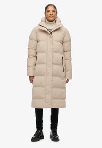 Beige gewatteerde winterjas met hoge kraag, lange lengte, voorrits en twee zijzakken. Heeft een capuchon en een gestructureerde afwerking.