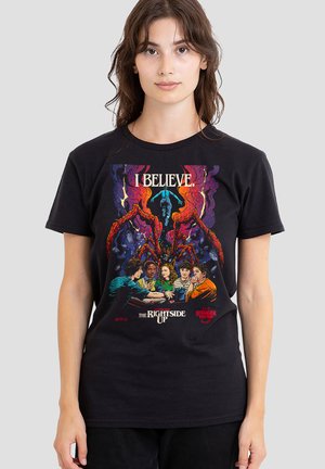 Woman wearing a black Stranger Things t-shirt featuring six characters at a table, a large red spider-like monster, and text "I BELIEVE."