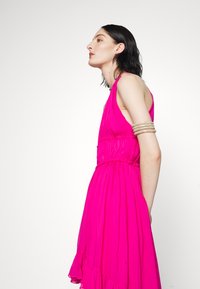 Vestito senza maniche di un rosa acceso, con texture plissettata e un design fluido. Presenta una vita arricciata e un orlo con balze. Accento di un bracciale d'argento.